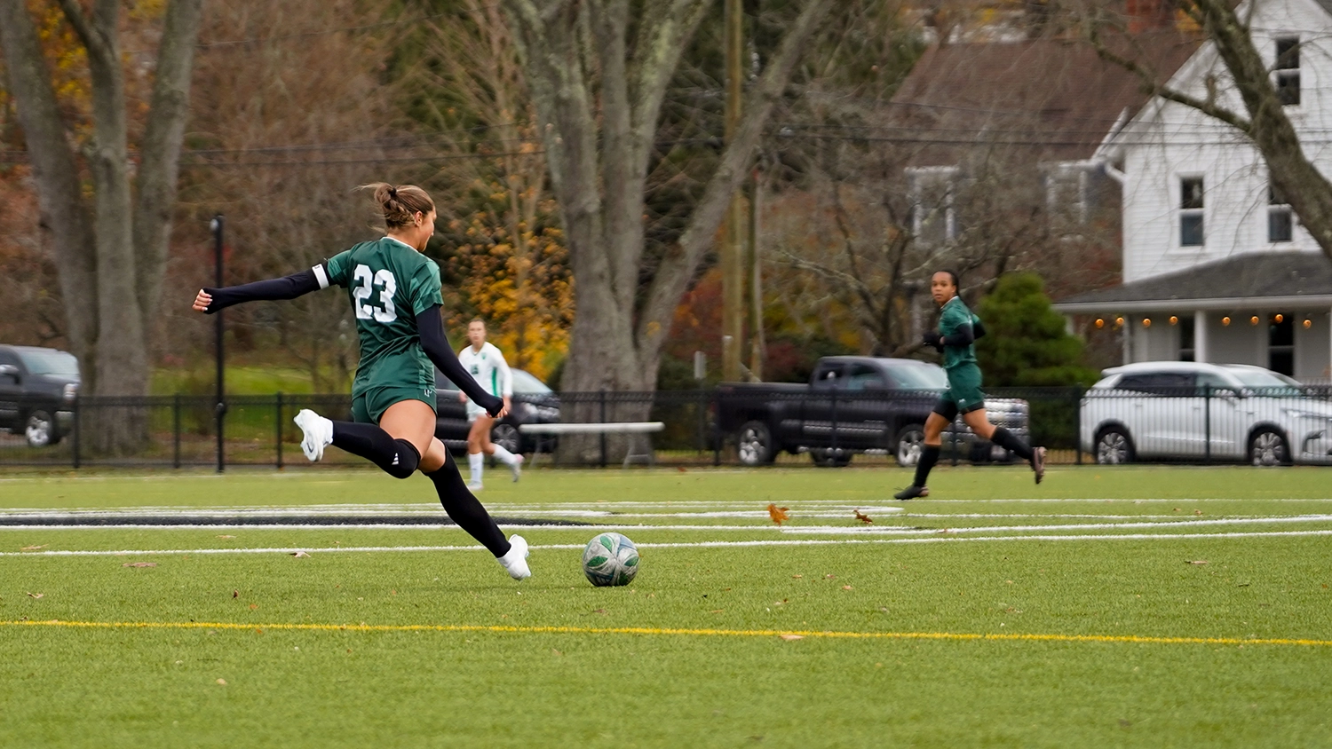 A soccer player in a green uniform kicks a ball on a field while teammates and a house appear in the background.