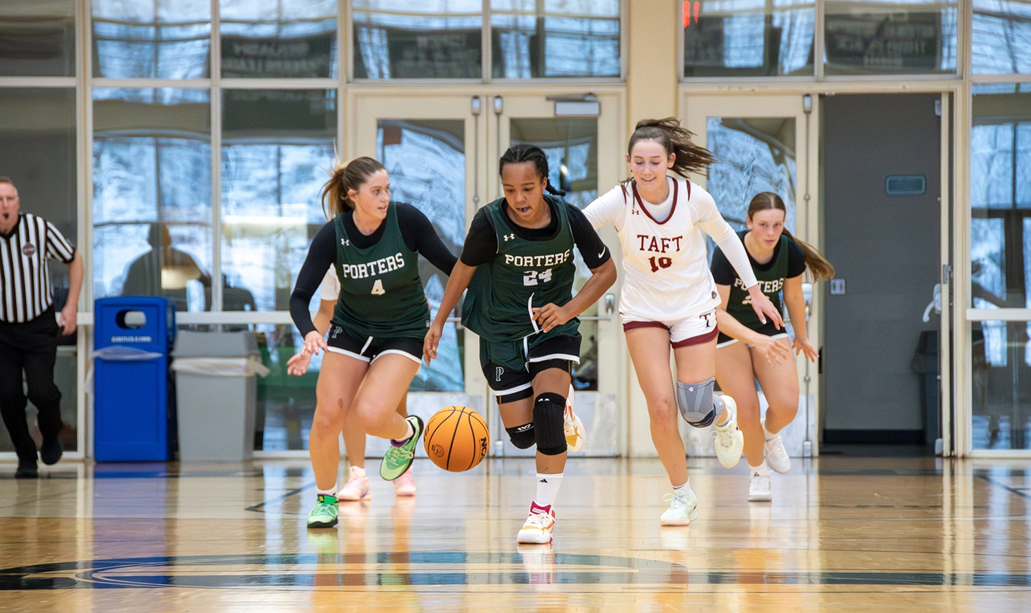Four high school basketball players chase a loose ball on an indoor court, with two players in green Porters uniforms and two in white Taft uniforms. A referee stands in the background.