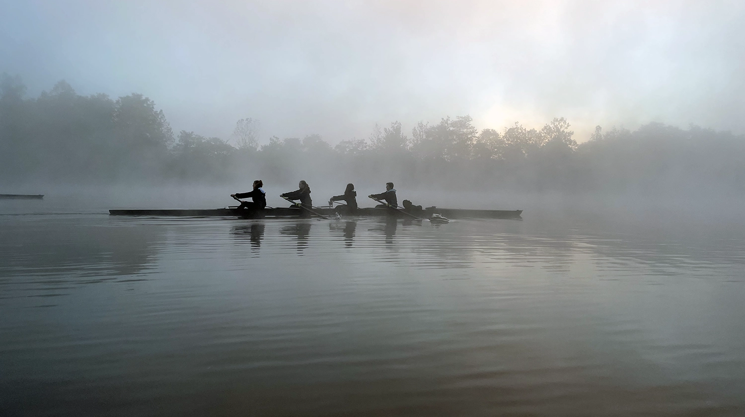 Four people row a long, narrow boat on a misty lake at dawn. Silhouetted against the fog, the rowers move in unison, with calm water and faint outlines of trees in the background.