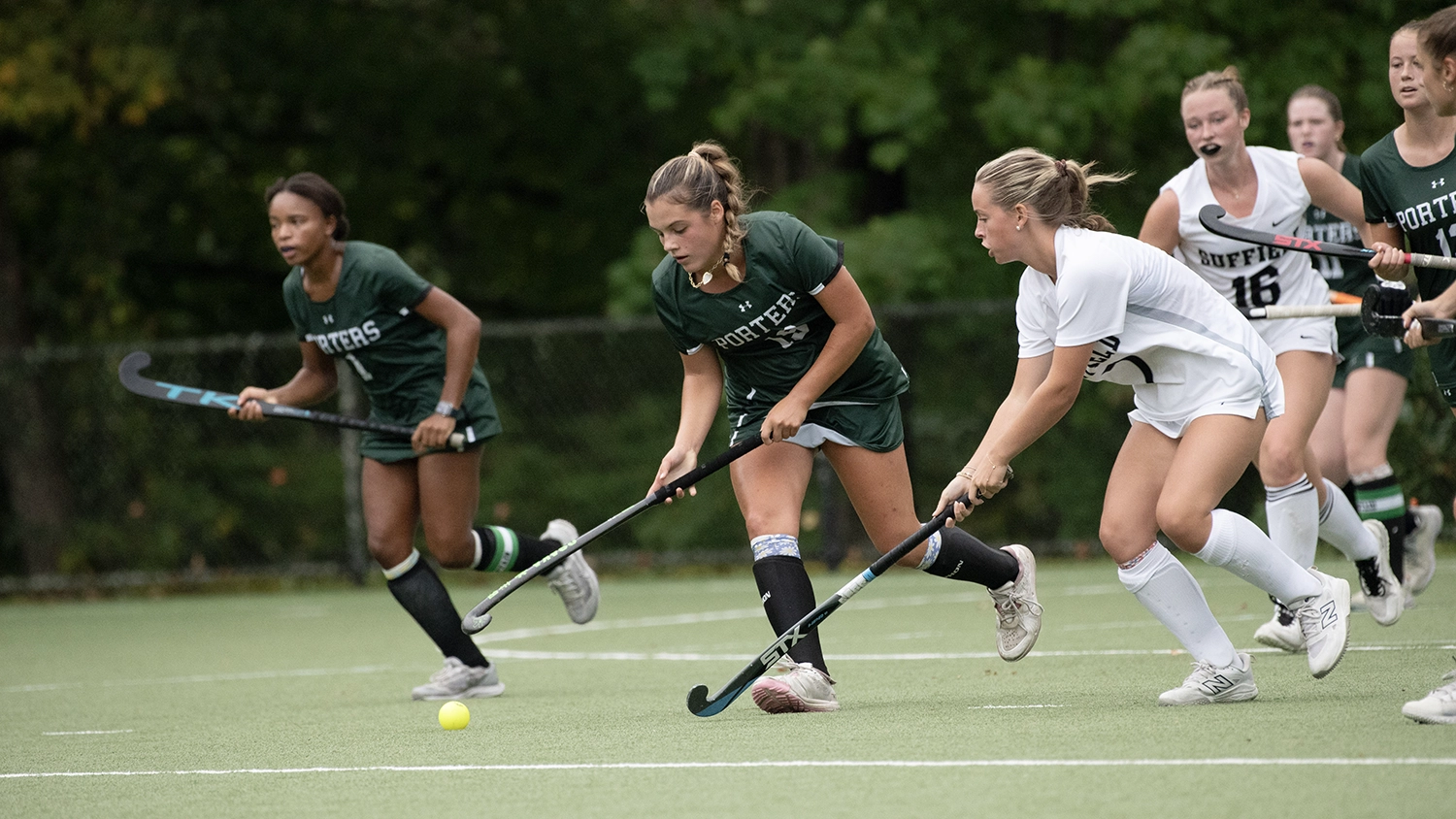 Four field hockey players chase a yellow ball on a green outdoor field, with trees in the background. Two wear green uniforms and two wear white, all focused and mid-action.