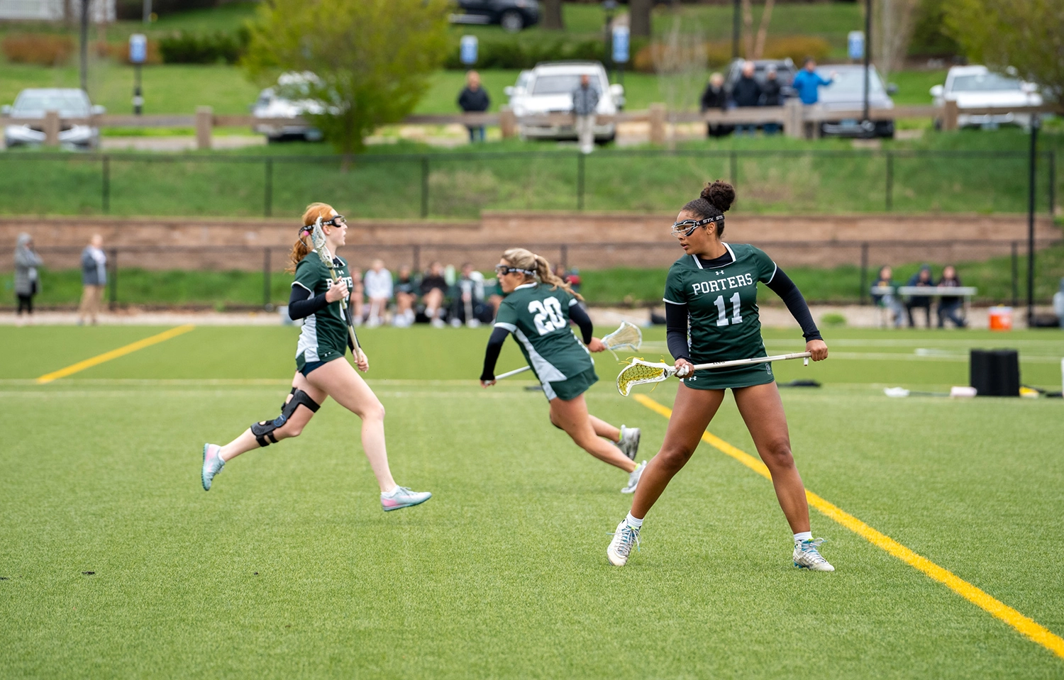 Four female lacrosse players in green uniforms run and play on a grassy field during a game, with spectators and parked cars visible in the background.