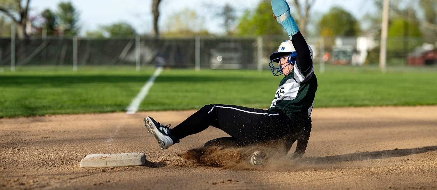 A female softball player wearing a helmet and face guard slides into a base, kicking up dirt on a sunny field. Trees and a fence are visible in the background.