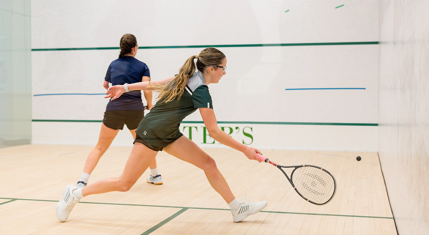 Two women play squash on an indoor court. One woman lunges forward with her racket to hit the ball, while the other stands behind her, ready for the next move. Both wear sports attire and protective eyewear.