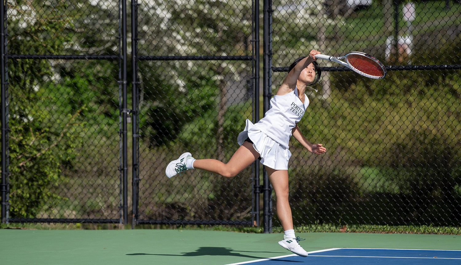A female tennis player in a white uniform jumps and reaches to hit a tennis ball on an outdoor court, with a black chain-link fence and greenery in the background.