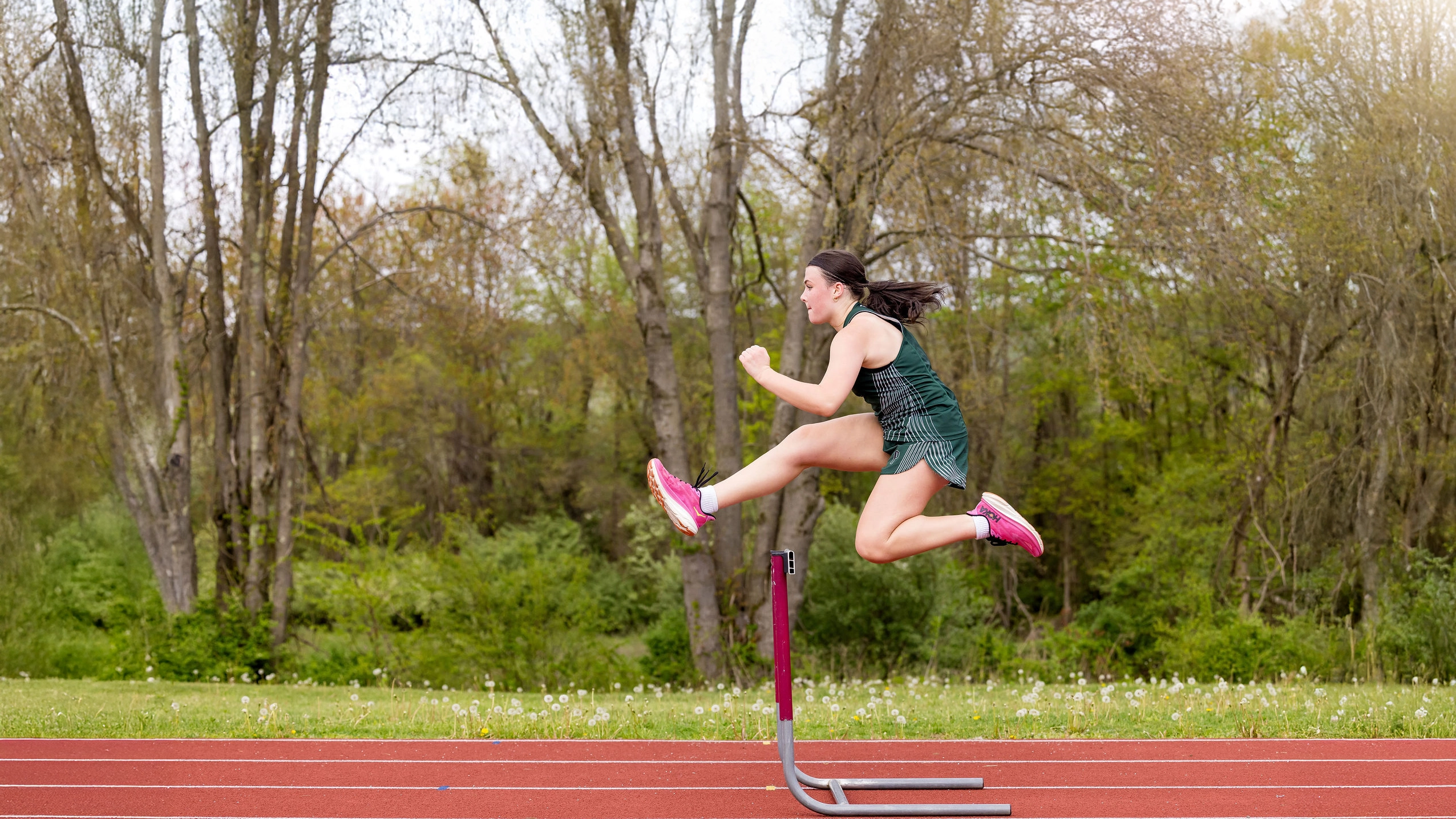 A female athlete in green sportswear and pink shoes jumps over a hurdle on an outdoor track, with trees and greenery in the background.