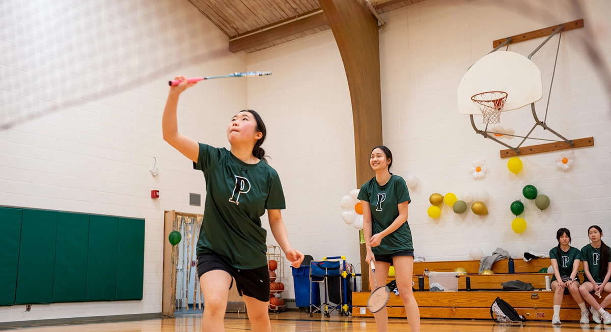Two girls in green shirts play badminton in a gym. One is about to hit the shuttlecock with her racket, while the other stands behind her. Three people sit on the bleachers in the background near a basketball hoop and balloons.