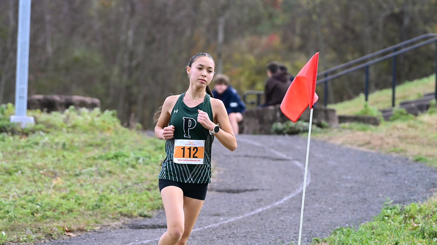 A young runner wearing a green jersey and race bib 112 runs on a trail near a red flag. The background shows grassy terrain, trees, and blurred people sitting on rocks.