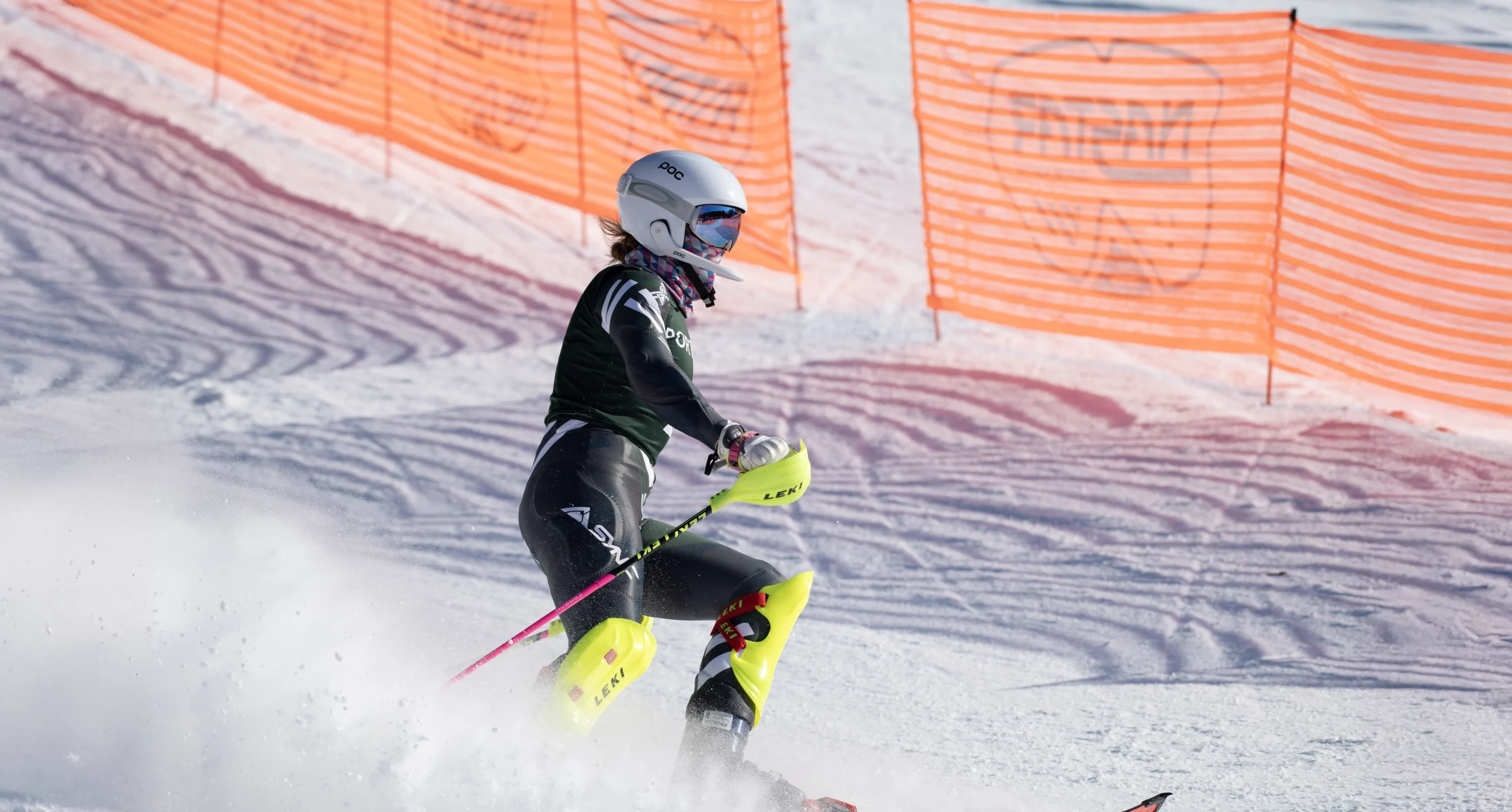 A skier in black and yellow gear, wearing a white helmet, navigates a snowy slope near orange safety netting. Snow flies up as they make a turn.