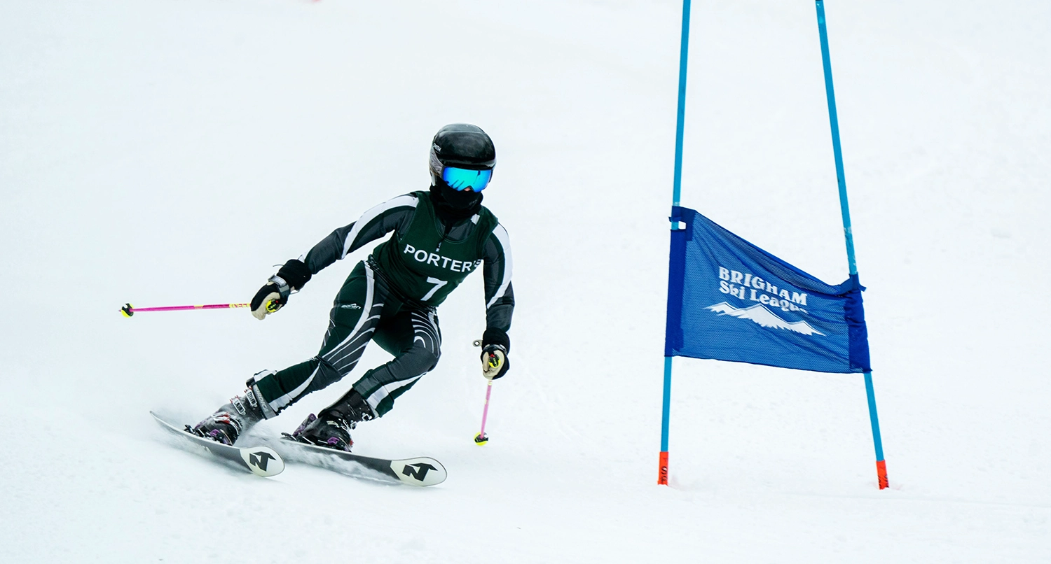 A skier in dark racing gear and helmet navigates around a blue slalom gate marked Brigham Ski Team on a snowy slope, holding ski poles and leaning into the turn.