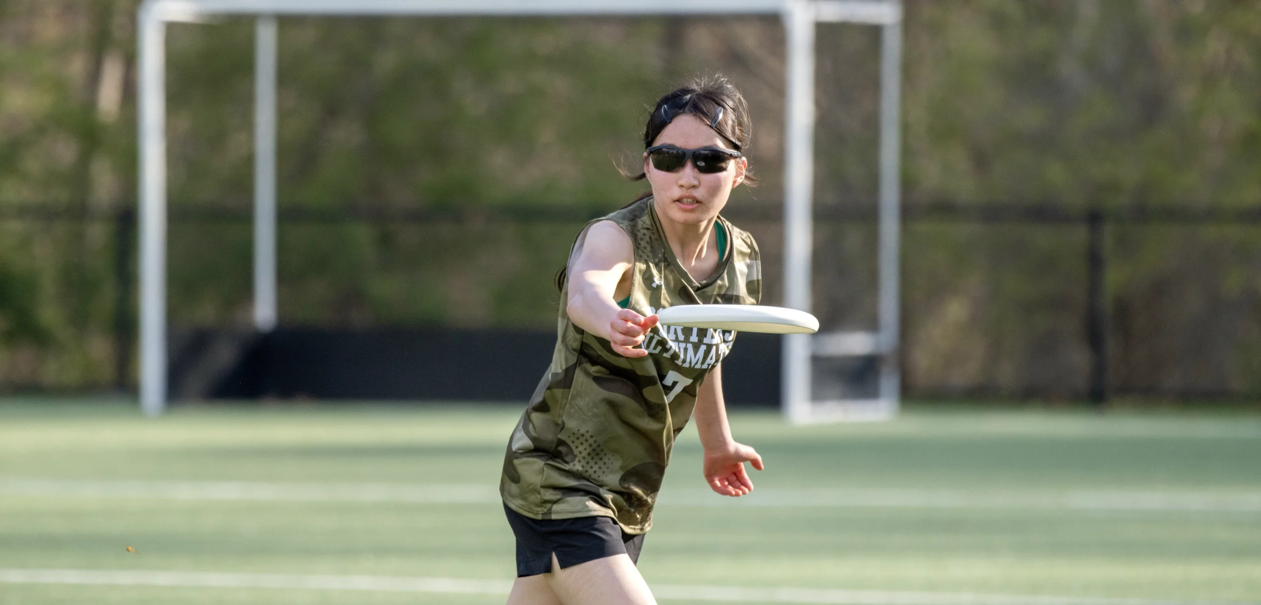 A person wearing sunglasses and a green sports jersey throws a frisbee on a grassy field with a soccer goal in the background.