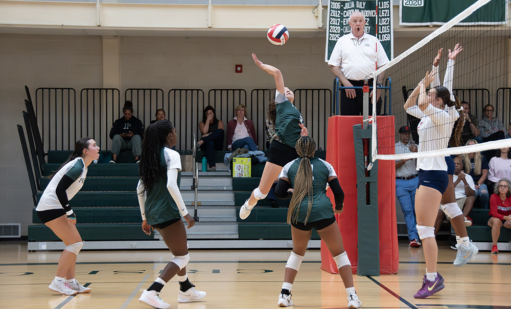 A Porter's volleyball player in a green jersey jumps to spike the ball over the net while teammates and opponents prepare to react; a referee observes from a stand, and spectators watch from the bleachers in the background.