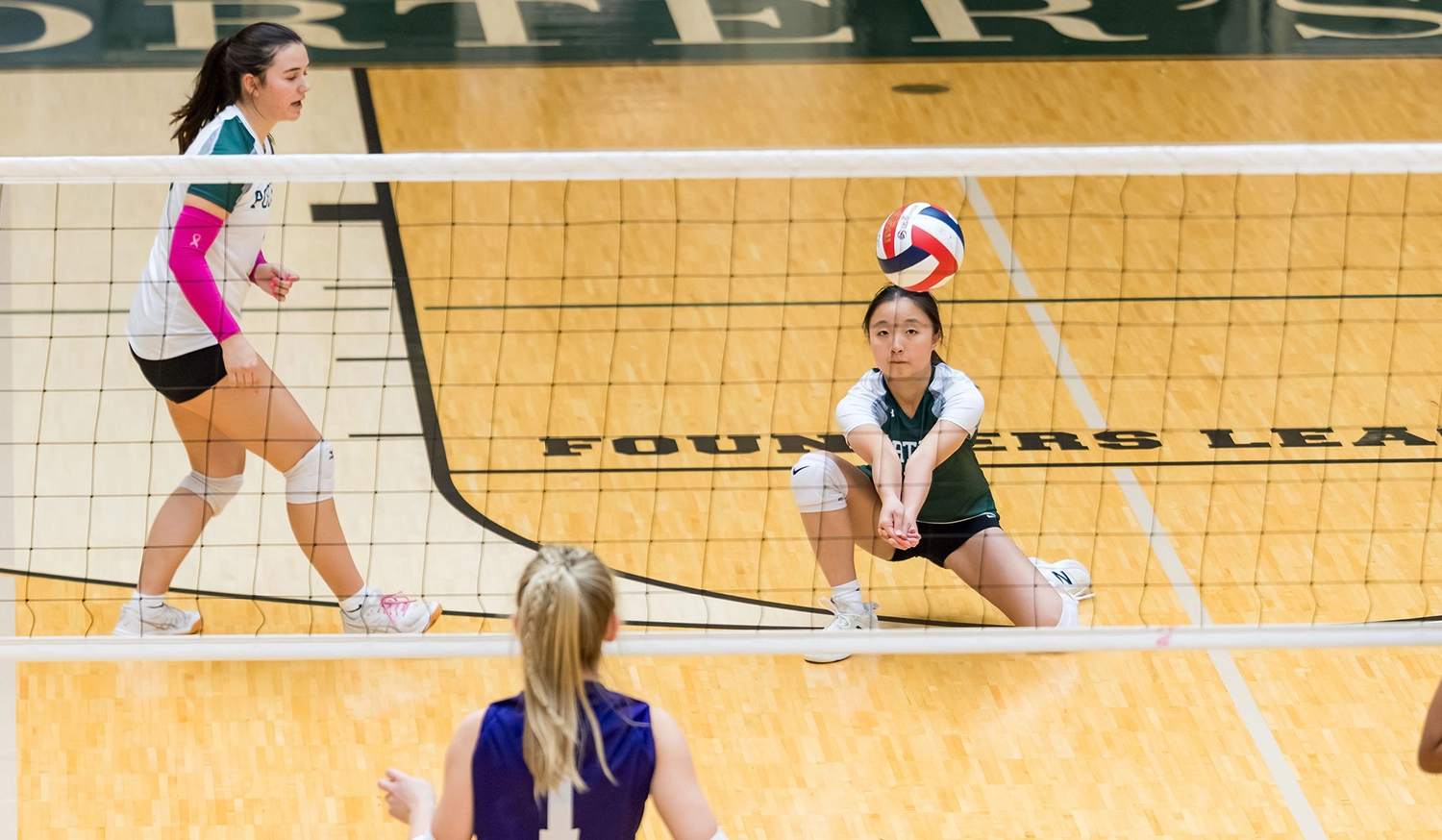A volleyball player in a green and white uniform crouches to bump the ball, while a teammate stands ready nearby. An opponent in a purple jersey stands at the net. The scene takes place on an indoor court.