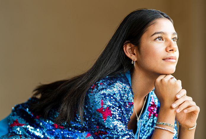 A young woman with long dark hair leans forward, resting her chin on her hands, gazing thoughtfully into the distance. She wears a blue sequined jacket with pink stars and several bracelets. The background is neutral.