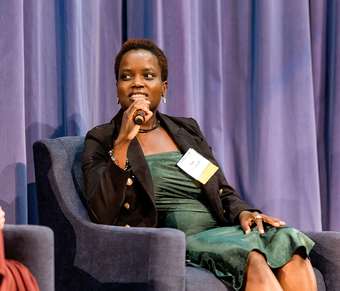 A woman wearing a black blazer and a green dress sits in a chair, speaking into a microphone. She has short hair and is wearing earrings and rings.
