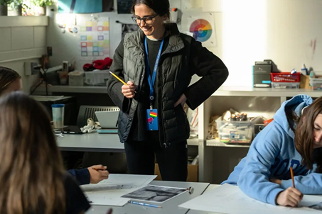 A teacher stands and smiles while interacting with students working on drawings at a classroom table filled with art supplies. Sunlight streams through a window, creating a bright and creative atmosphere.