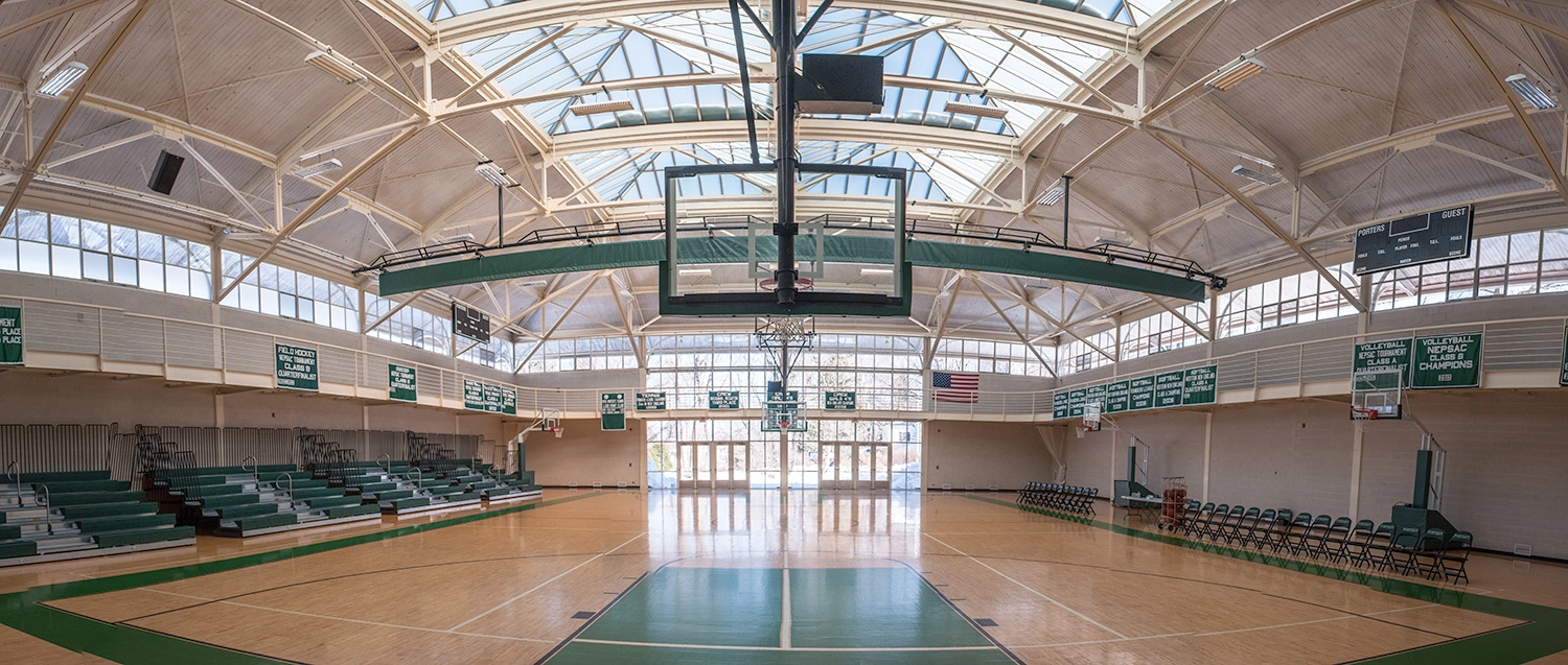 A spacious indoor basketball gym with a high, arched ceiling, large windows, empty bleachers, a scoreboard, and an American flag on the wall. The polished court is lit by natural light streaming through skylights.
