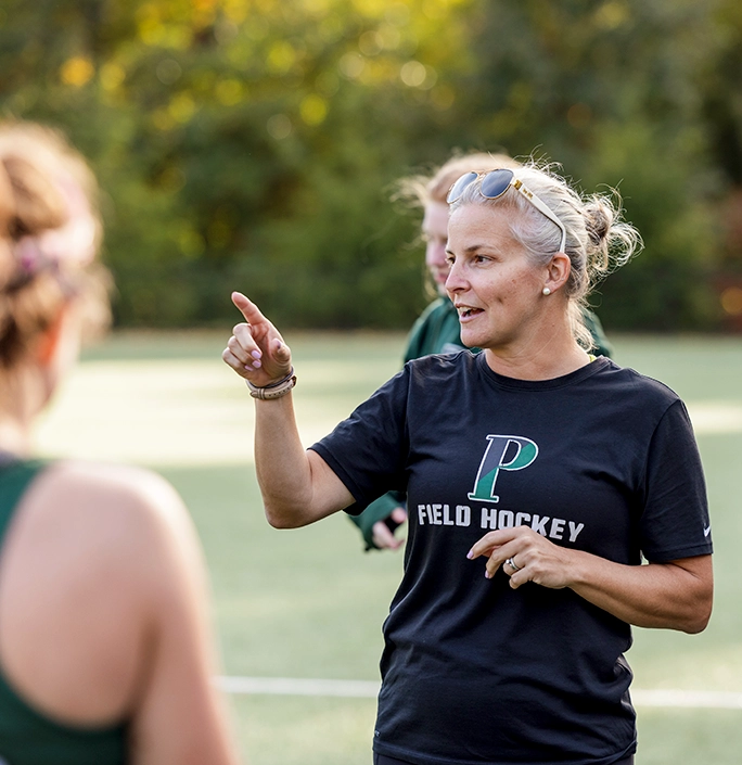A woman wearing sunglasses on her head and a P Field Hockey shirt gestures while speaking to players on a field during daylight, with trees blurred in the background.