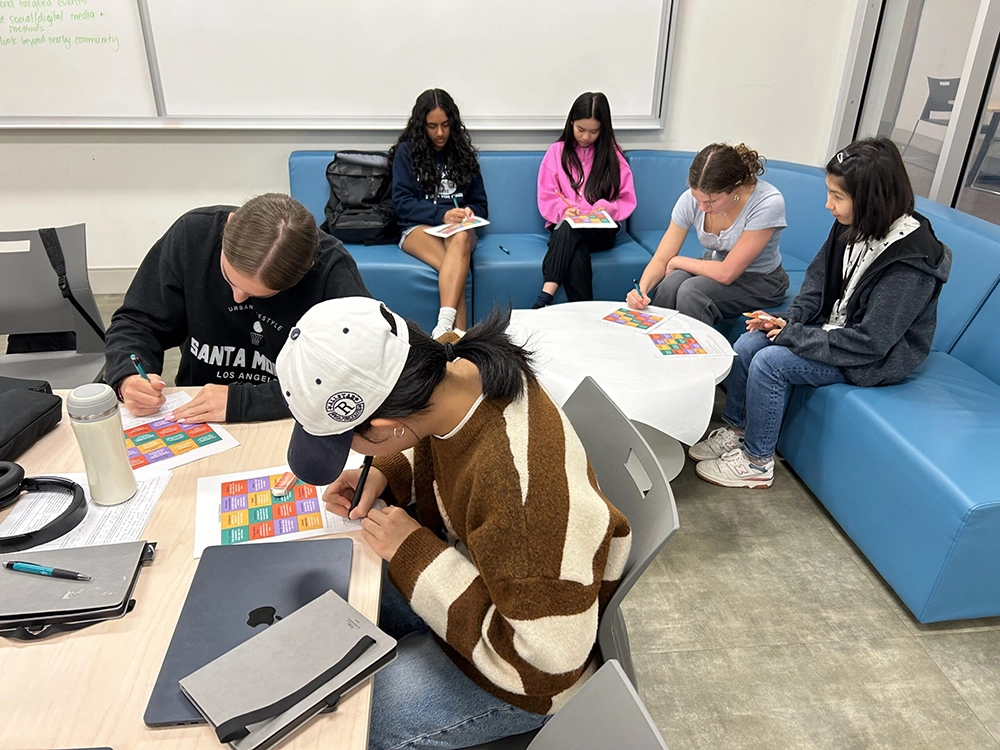 Six students are seated in a classroom, writing or filling out colorful charts at tables and couches, with notebooks, laptops, and water bottles also visible.