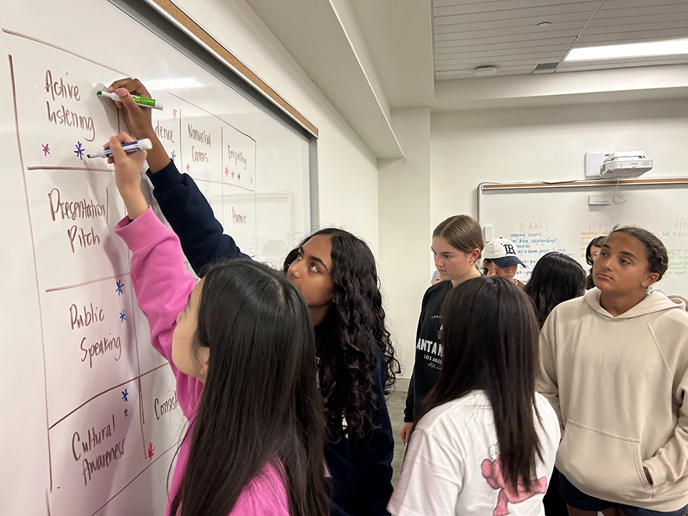 A group of students stands by a whiteboard, where one student is writing under the Active Listening category while others watch and discuss. The classroom setting appears collaborative and focused.