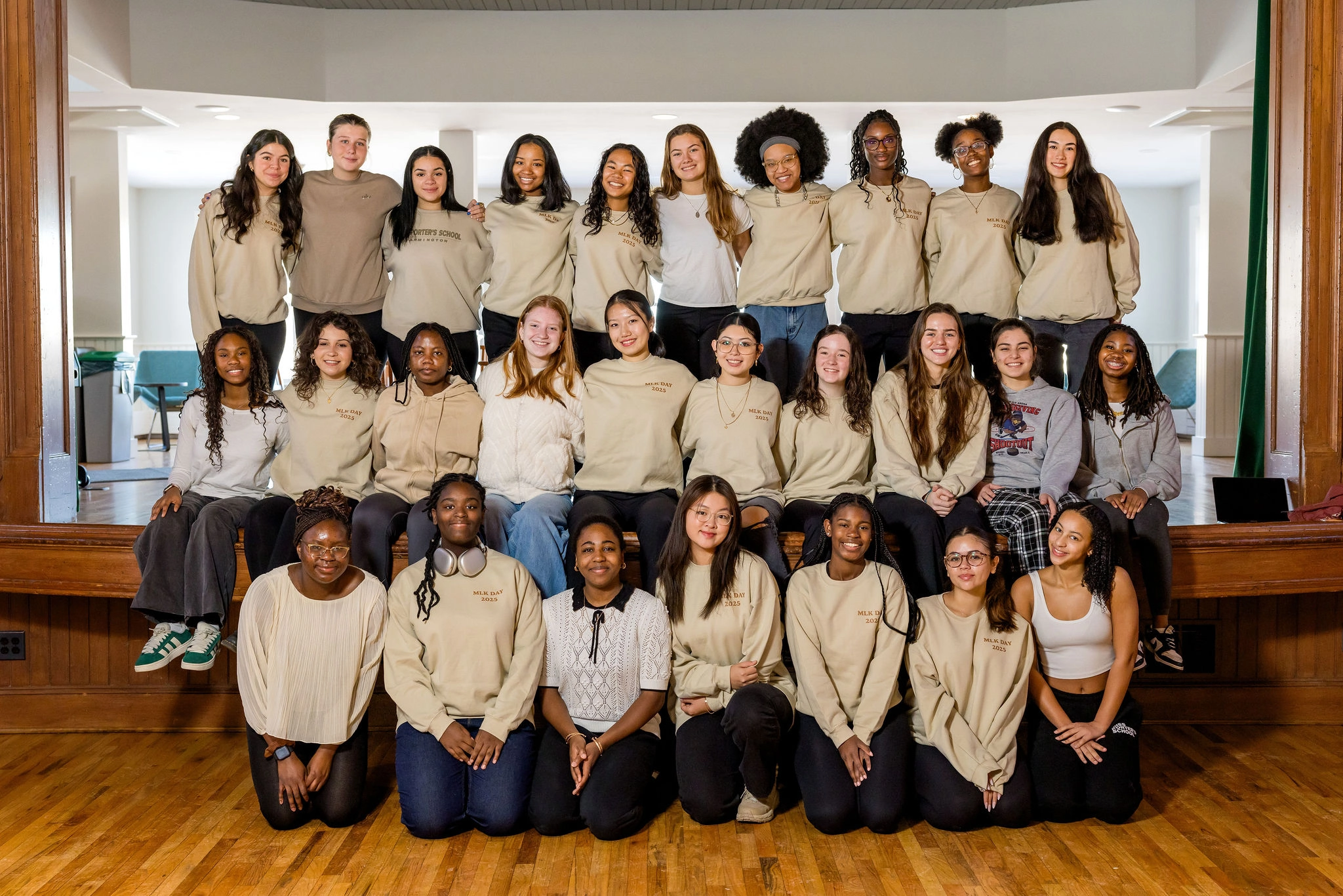 A group of 27 diverse young women pose together on a stage. Most wear matching beige sweatshirts, and they are arranged in four rows, smiling and looking at the camera in a bright indoor setting.