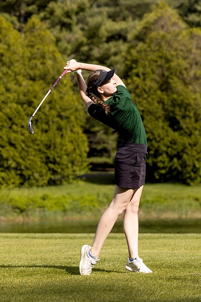 A young woman wearing a green shirt, black shorts, and a visor swings a golf club on a sunny golf course with trees in the background.