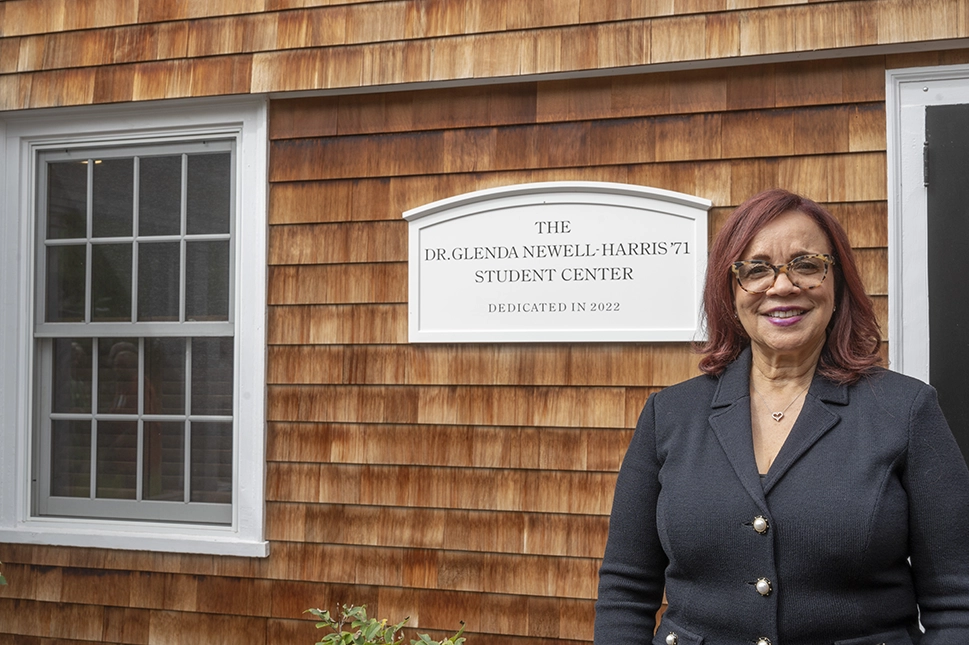 A woman with red hair and glasses stands smiling in front of a building with a sign that reads, The Dr. Glenda Newell-Harris 71 Student Center Dedicated in 2022.