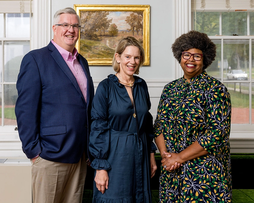 Three adults stand indoors, smiling at the camera. A man in a blue blazer and pink shirt stands next to two women, one in a dark dress and the other in a colorful patterned dress. A framed painting hangs on the wall behind them.