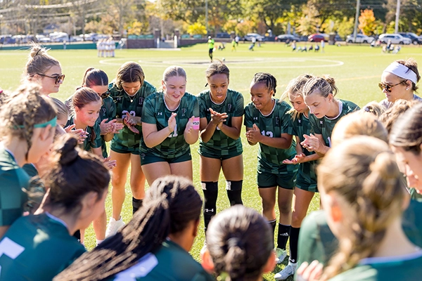 A girls soccer team, wearing green uniforms, stands in a huddle on a sunny field, smiling and clapping together before a game. Trees and another team are visible in the background.