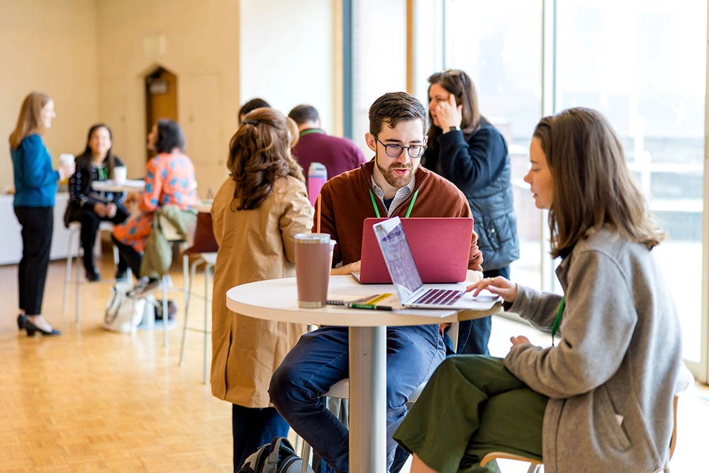 People sit and stand around tables in a bright room, talking and working on laptops. The atmosphere is casual and collaborative, with large windows letting in natural light—ideal for exploring opportunities for educators.