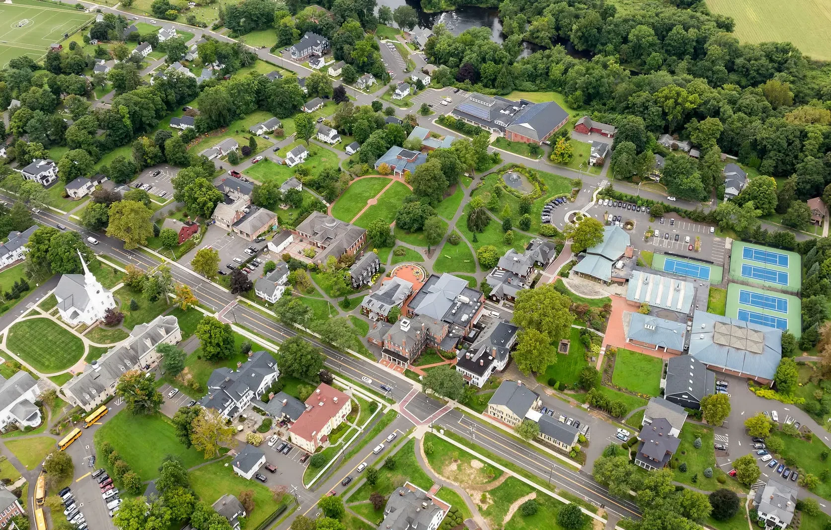 Aerial view of Miss Porter's School campus with school buildings, houses, roads, a church, parking lots, tennis courts, and green trees surrounding the area. Fields and wooded areas are visible in the background.