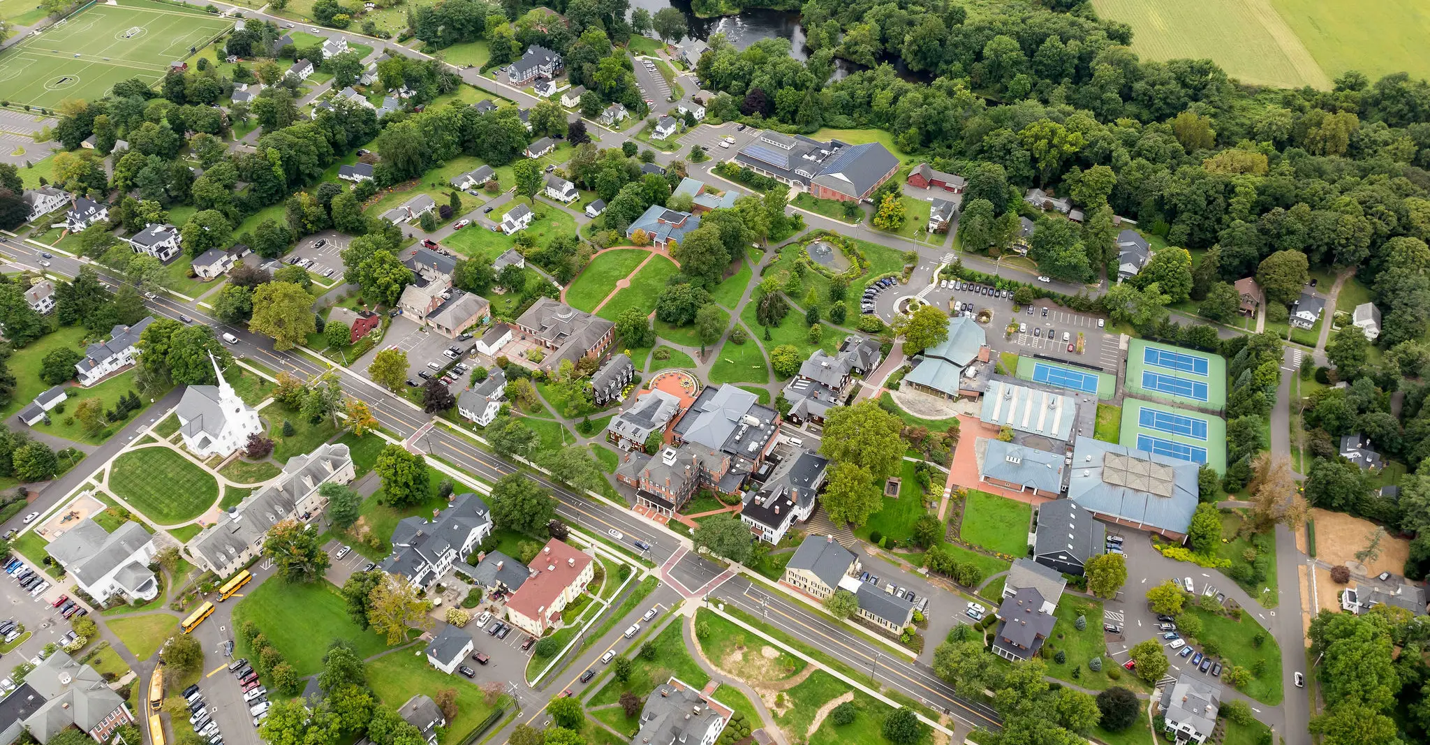 Aerial view of Miss Porter's School campus with school buildings, houses, roads, a church, parking lots, tennis courts, and green trees surrounding the area. Fields and wooded areas are visible in the background.