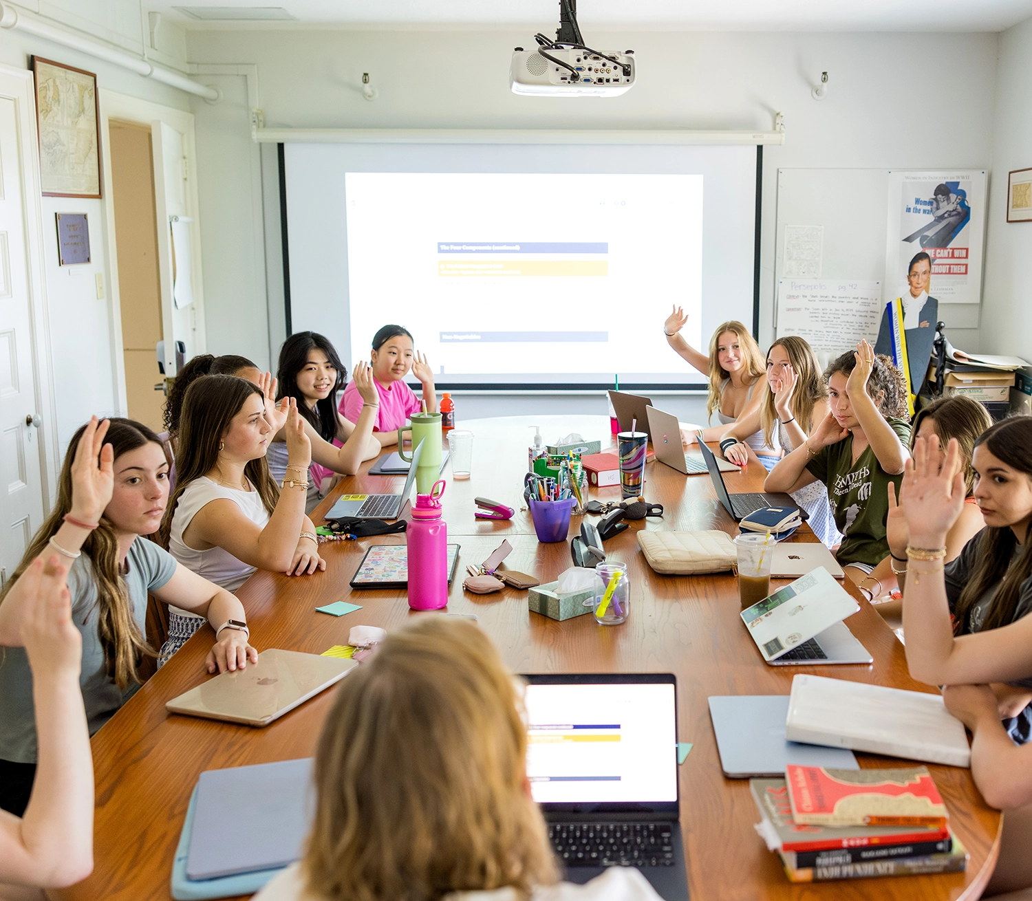 A group of students sit around a conference table with laptops and notebooks, raising their hands in a classroom with a large projection screen at the front.