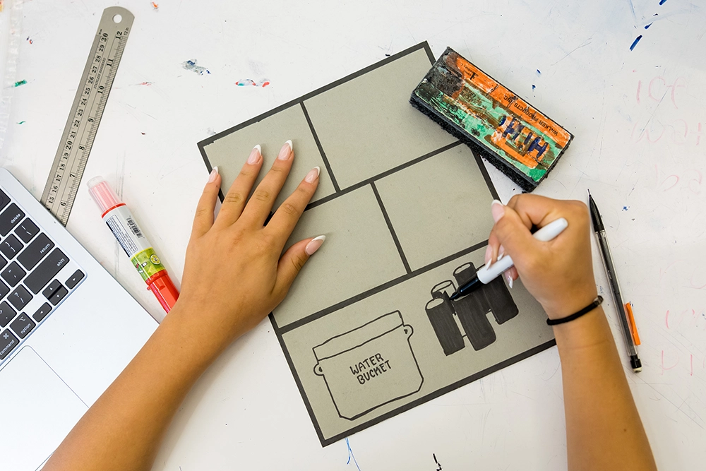 A person draws on a gray comic panel page, outlining objects including a bucket labeled WATER BUCKET. Art supplies, a laptop, and a ruler are on the white desk.