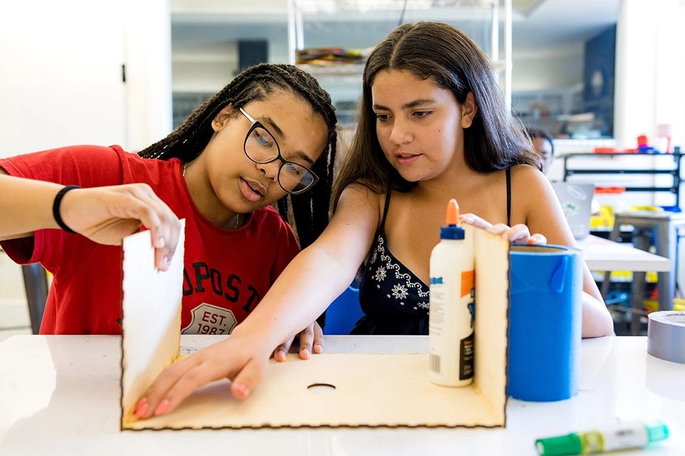 Two girls work together at a table, assembling a wooden project using glue. Craft supplies like tape and a blue container are on the table in a bright, modern classroom.