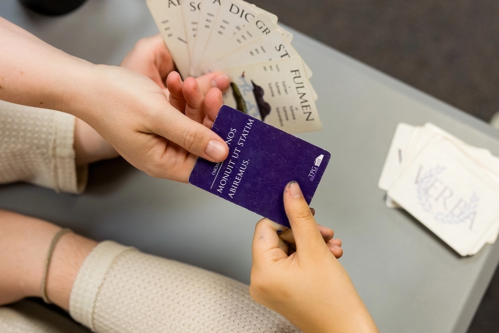 Two girls play a card game; one person hands a purple card with Latin text to another, who is wearing a light-colored, long-sleeve shirt and a smartwatch. Cards with Latin words and letters are visible.