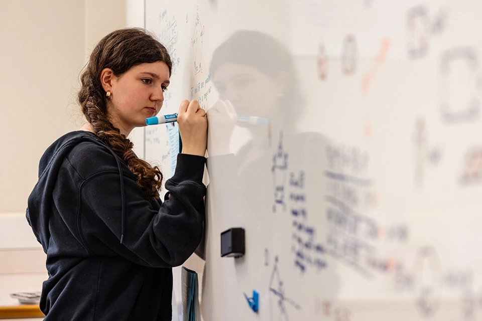 A young woman with long braided hair writes on a whiteboard filled with colorful notes, diagrams, and sketches in a classroom or study setting.