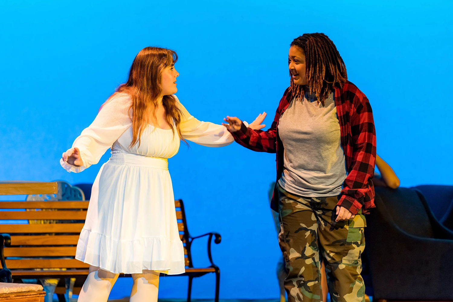 Two people perform onstage. One wears a white dress and gestures strongly, while the other, dressed in a red plaid shirt and camouflage pants, responds with a raised hand. A bench and blue backdrop are visible behind them.