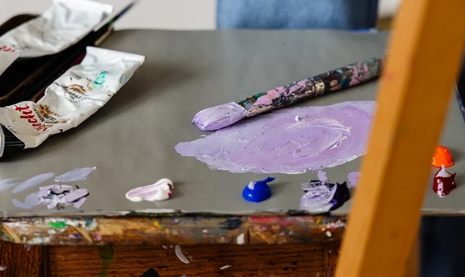 A close-up of a painter’s palette with mixed purple paint, blobs of blue, red, orange, and white, a paintbrush, and paint tubes on a wooden easel. A person in jeans stands in the background.