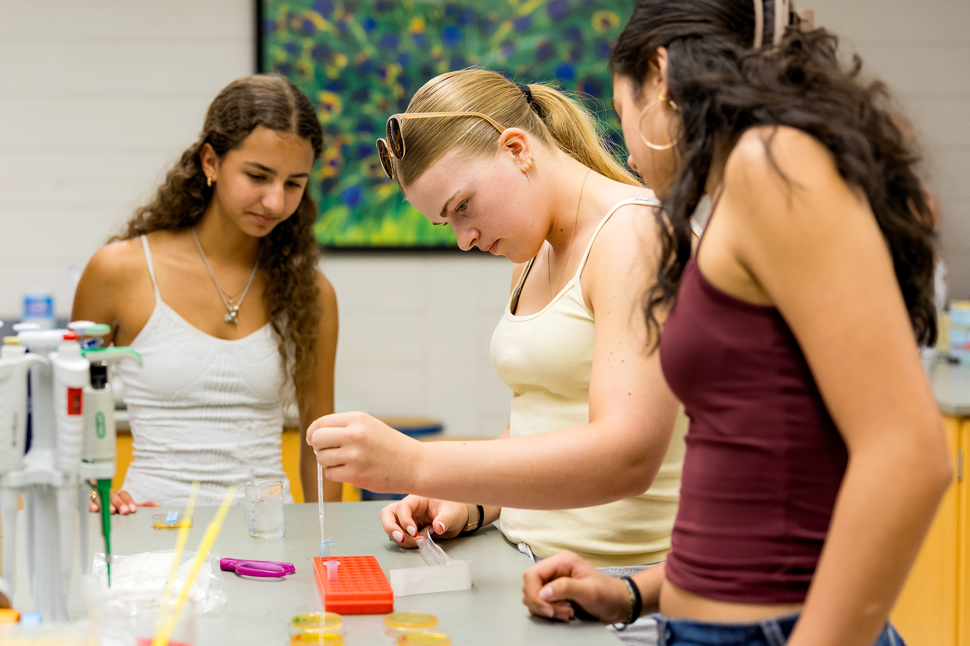 Three young women in a science lab work together with pipettes and petri dishes, focusing on an experiment at a lab bench.