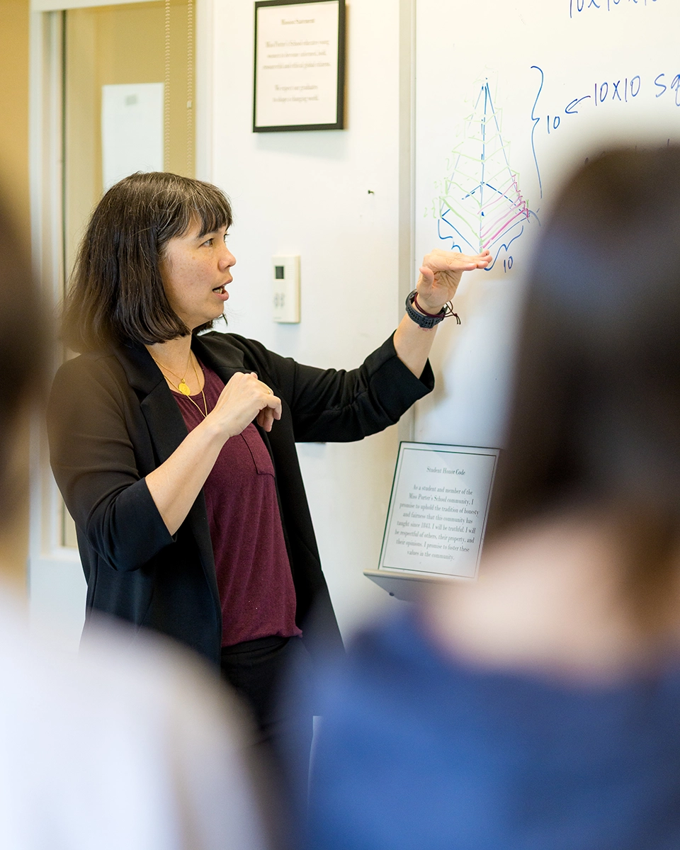 A teacher in a black blazer and maroon top stands before a whiteboard, gesturing toward a drawn geometric pyramid diagram with labels. Students are visible in the foreground out of focus. A posted honor code is on the wall behind her.