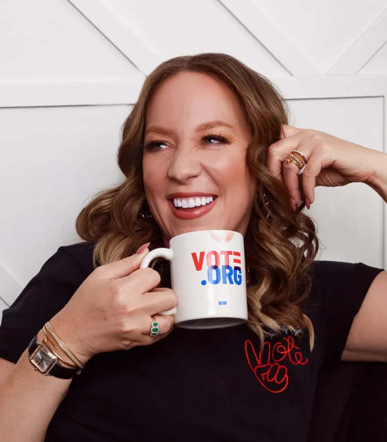 A woman with wavy light brown hair smiles and holds a white mug that says VOTE.ORG in red and blue letters. She wears a black shirt with a red heart and gold jewelry, sitting against a white wall.