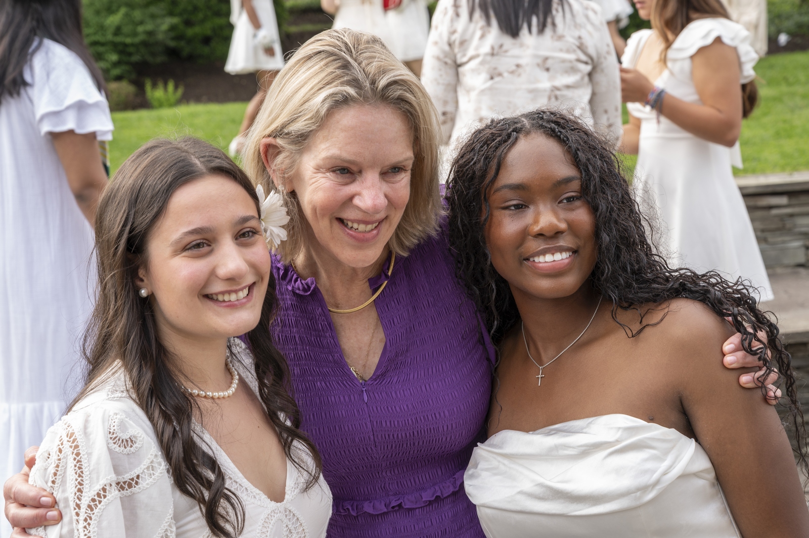 Three women pose and smile outdoors. Two wear white dresses, and one in the middle wears a purple dress with short sleeves. Other people in white outfits are visible in the background on a grassy area.