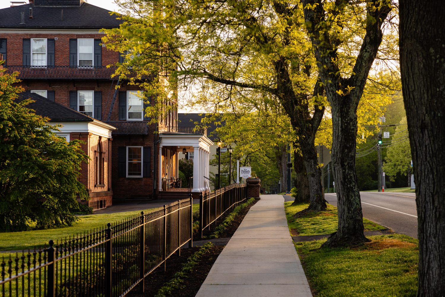 A sidewalk lined with trees runs alongside a wrought iron fence and a brick building at sunset, with soft golden light filtering through the green leaves.