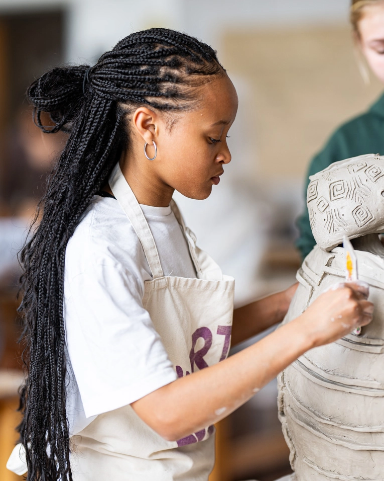 A woman with long braids, wearing hoop earrings and an apron, carefully sculpts and paints a large clay pottery piece in a studio setting.