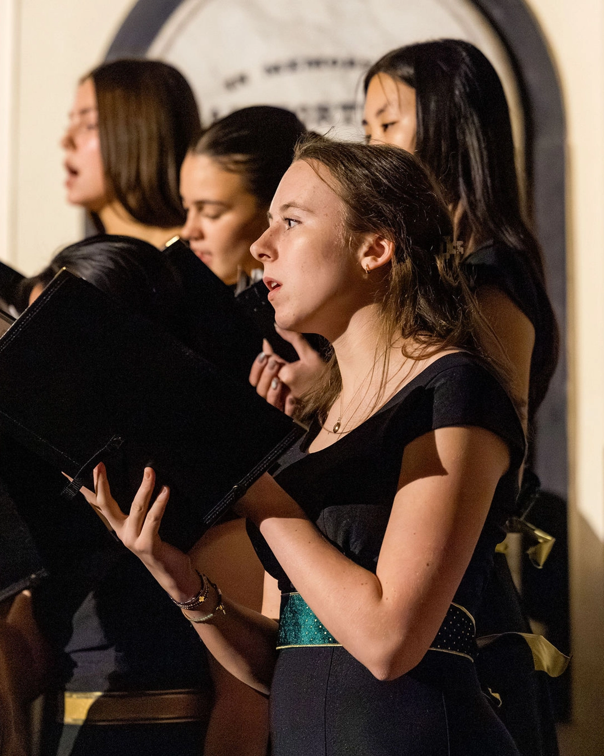 A group of young women are singing in a choir, each holding black folders with sheet music. The lighting highlights one singer in the foreground, who appears focused and expressive.