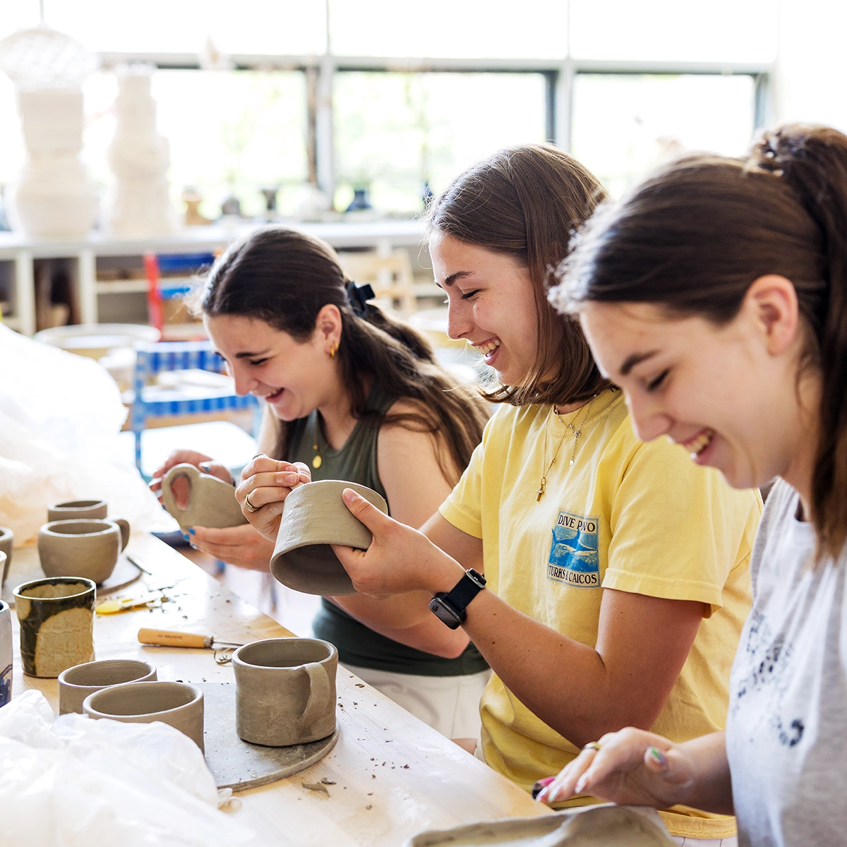 Three young women sit at a table in a bright studio, smiling and shaping clay mugs with pottery tools. Various clay pieces and supplies are on the table, and shelves are visible in the background.