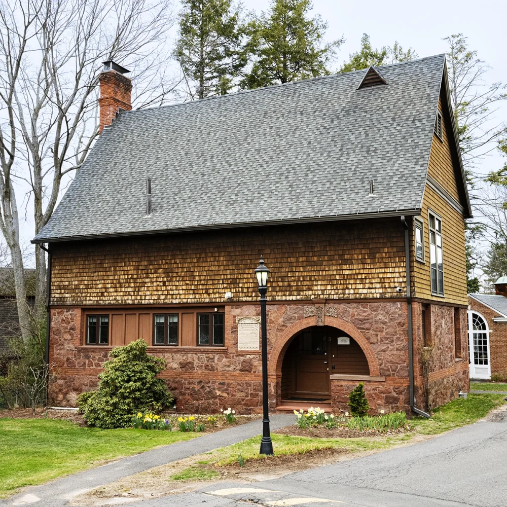 A brown stone and shingle house with a steep roof, arched doorway, and a streetlamp in front, surrounded by greenery and yellow flowers. Trees and sky are visible in the background.