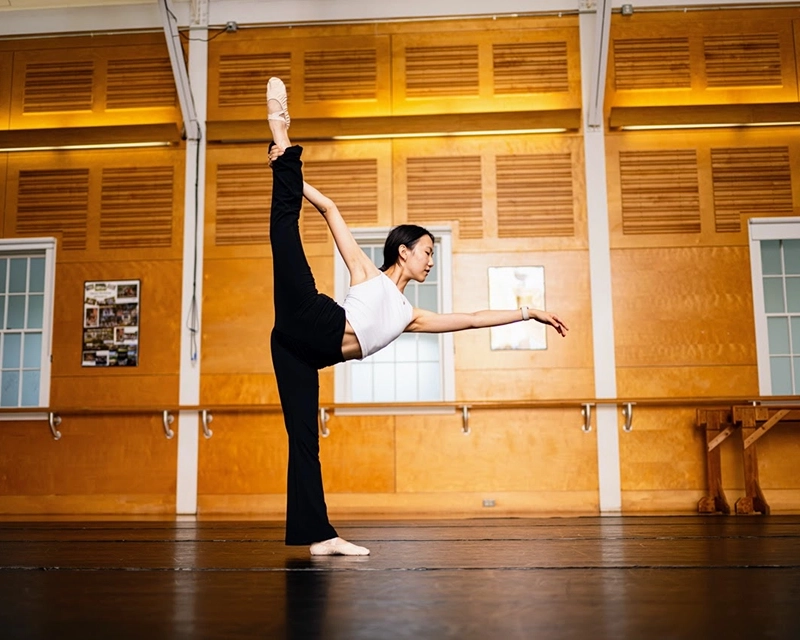 A dancer in a studio balances on one leg while holding the other leg extended high behind her, wearing a white top and black pants. The studio has wood-paneled walls and large windows.