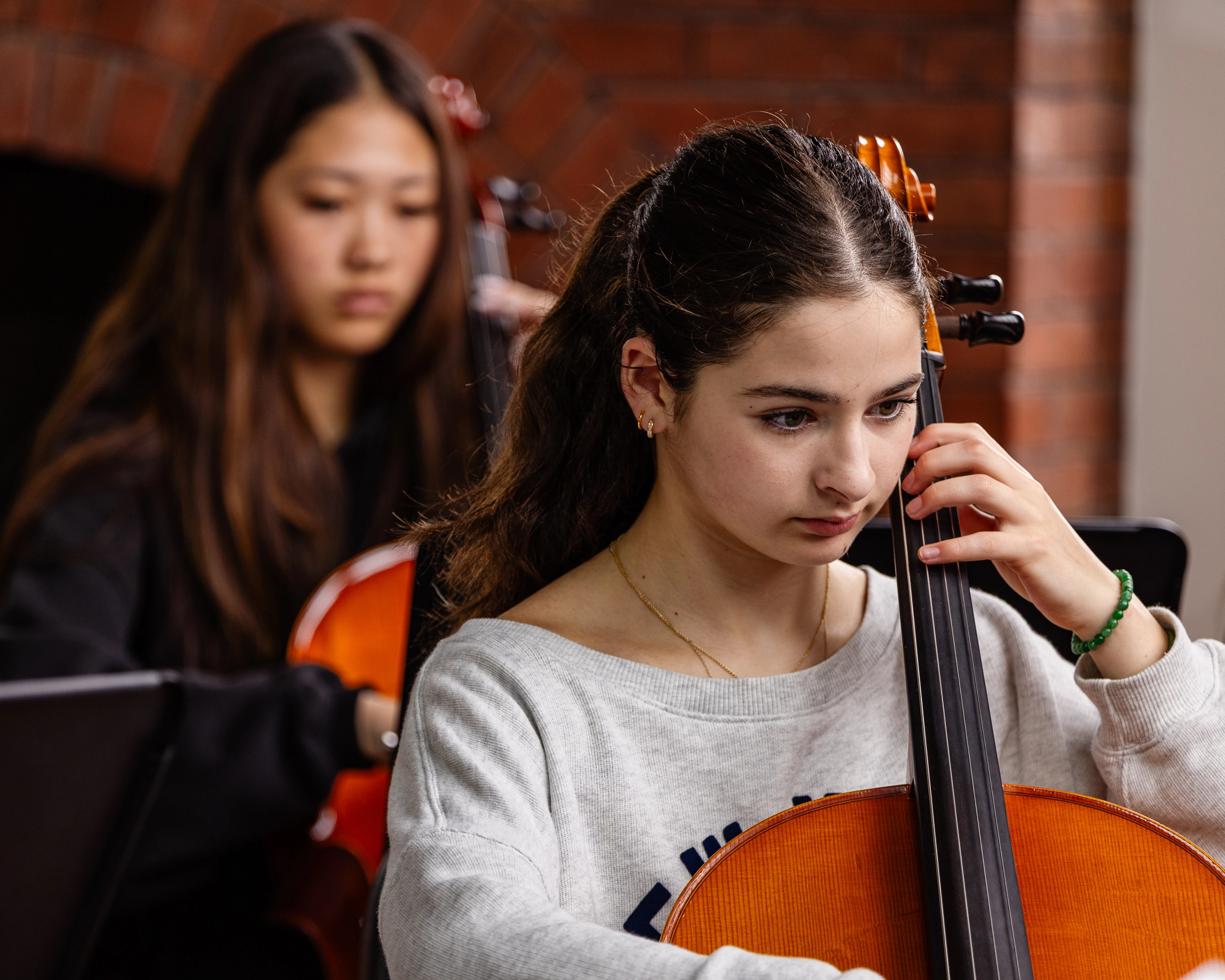 Two young women play cellos indoors. The focus is on the woman in front with dark brown hair and a light gray sweater; the woman behind her is blurred. Both appear concentrated on their music. Brick wall in the background.