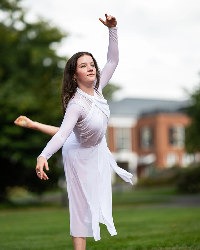 A young woman in a white dress performs a graceful dance pose outdoors, one arm raised and one leg extended behind her, with a building and trees in the background.